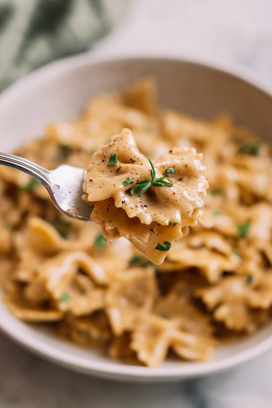 A close-up view shows a white bowl filled with creamy pasta that has a light brown sauce, evenly coating the pasta with small green herbs sprinkled on top. In the foreground, a silver fork holds a small portion of the wavy-edged pasta covered in sauce with visible pepper specks and tiny green leaves. The background is softly blurred, showing more pasta in the bowl resting on a white marbled surface. The photo taken with an iphone --ar 2:3 --v 7 - French Onion Pasta, French Onion Soup Pasta, Caramelized Onion Pasta, Cozy Pasta Recipes, Easy Pasta Dinner