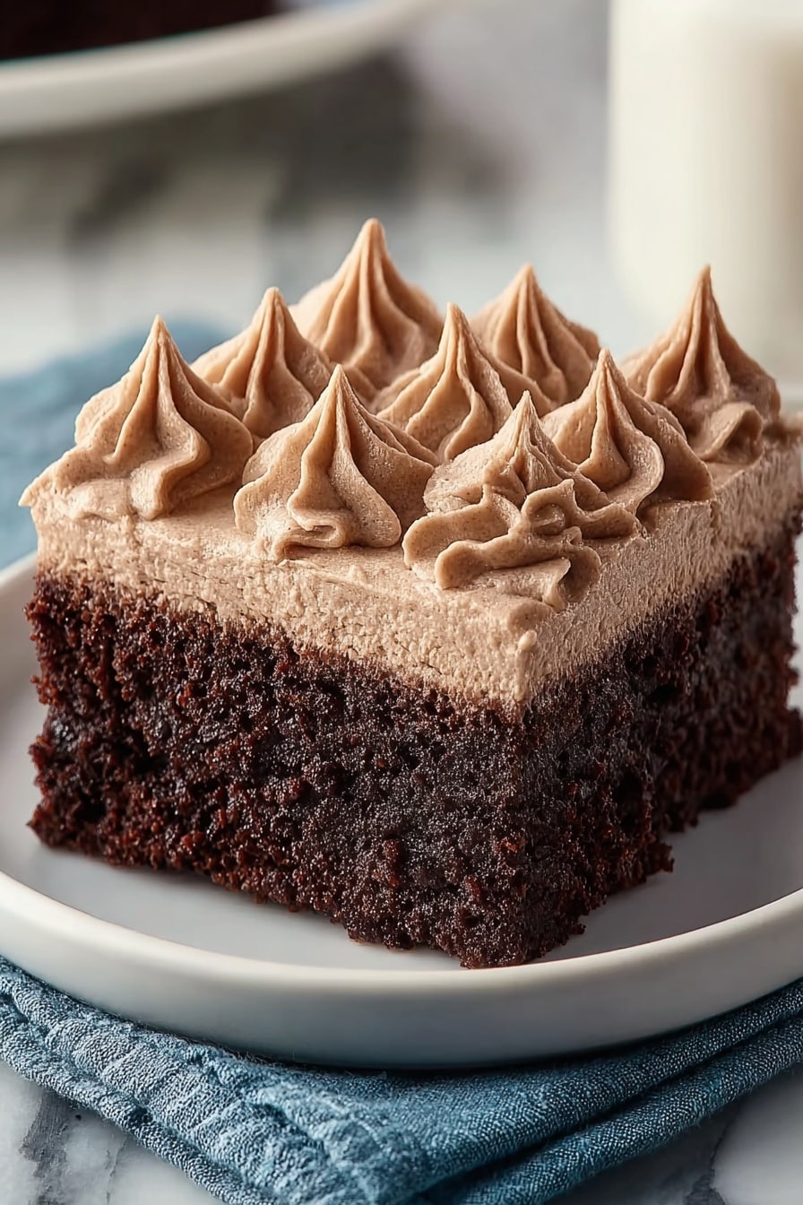 A square piece of chocolate cake with two layers sits on a white plate placed on a blue cloth and white marbled surface. The bottom layer is dark brown and has a moist, slightly crumbly texture. The top layer is a smooth, light brown frosting that is thicker than the cake part. On top of the frosting, there are ten small peaks of the same light brown frosting, piped in a swirl pattern to add texture and height to the piece. Photo taken with an iphone --ar 2:3 --v 7 - Frosted Coffee Brownies, coffee-flavored brownies, chocolate coffee dessert, easy coffee brownies, chocolate brownie recipes