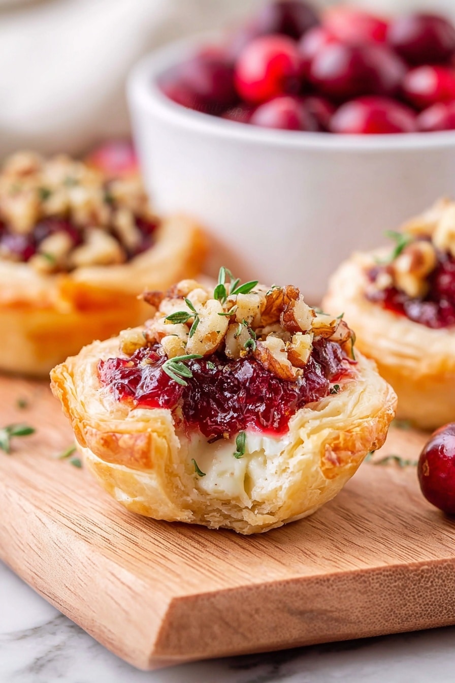 The image shows a close-up of a small tart with three visible layers: a golden, flaky puff pastry shell on the outside, a middle layer of melted creamy cheese with a smooth texture, and a top layer of bright red cranberry sauce mixed with chopped nuts and garnished with small green herb leaves. The tart sits on a light wooden board, with another tart and a white bowl filled with red cranberries blurred in the background, all placed on a white marbled surface. photo taken with an iphone --ar 2:3 --v 7 - Cranberry Brie Bites, Cranberry Brie Bites appetizer, easy cranberry brie bites, festive puff pastry appetizers, holiday cranberry brie bites