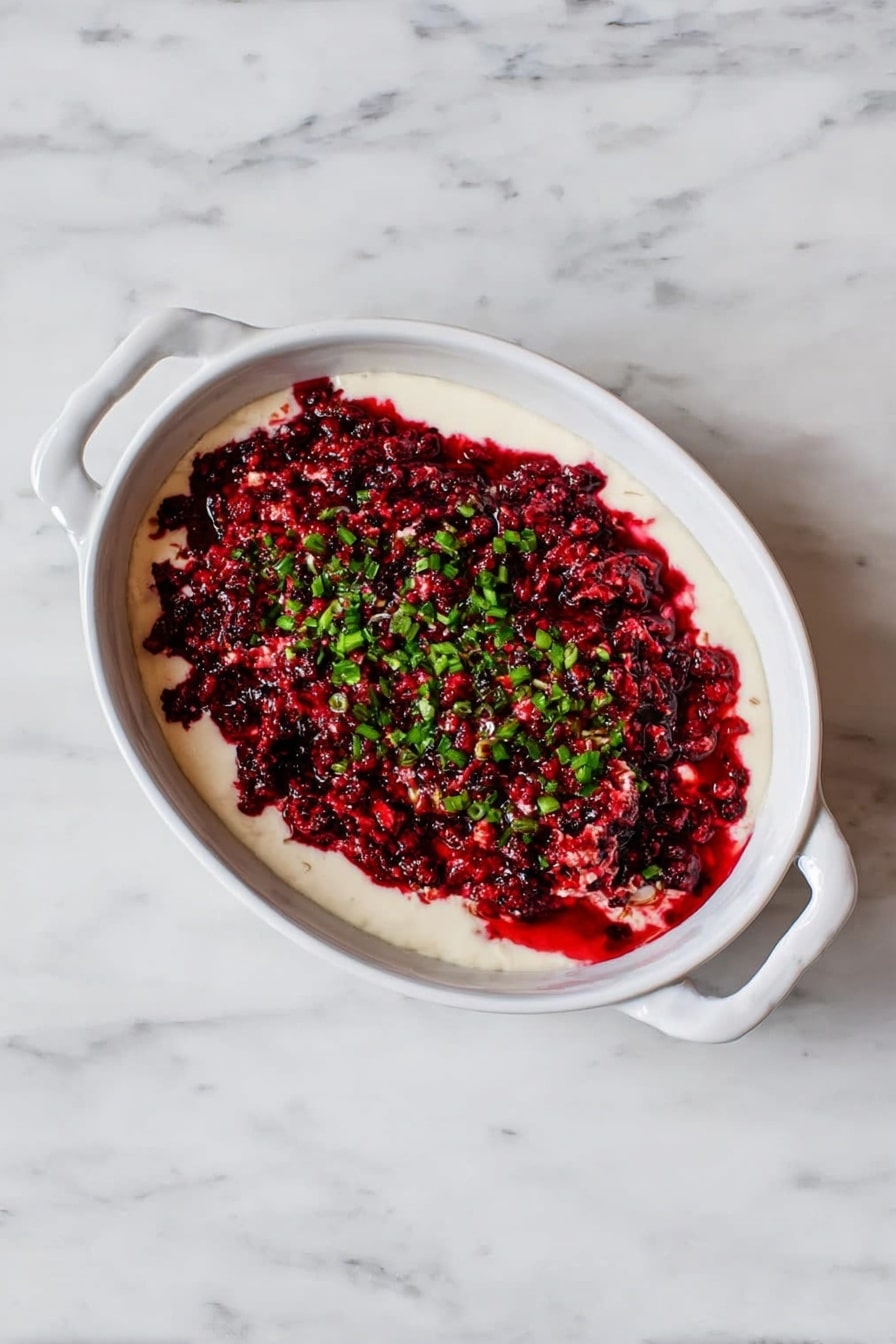 The image shows a close-up of a white plate with a textured soft white creamy spread as the bottom layer. On top of this spread is a thick layer of finely chopped red and pink mixed fruit or berries, mixed with small pieces of green herbs scattered evenly across the surface. A woman's hand is holding a light beige chip dipped in the red mixture, lifted near the edge of the plate. The plate rests on a white marbled surface. photo taken with an iphone --ar 2:3 --v 7 - Cranberry Jalapeño Cream Cheese Dip, spicy cranberry dip, holiday appetizer, easy party dip, creamy fruit dip
