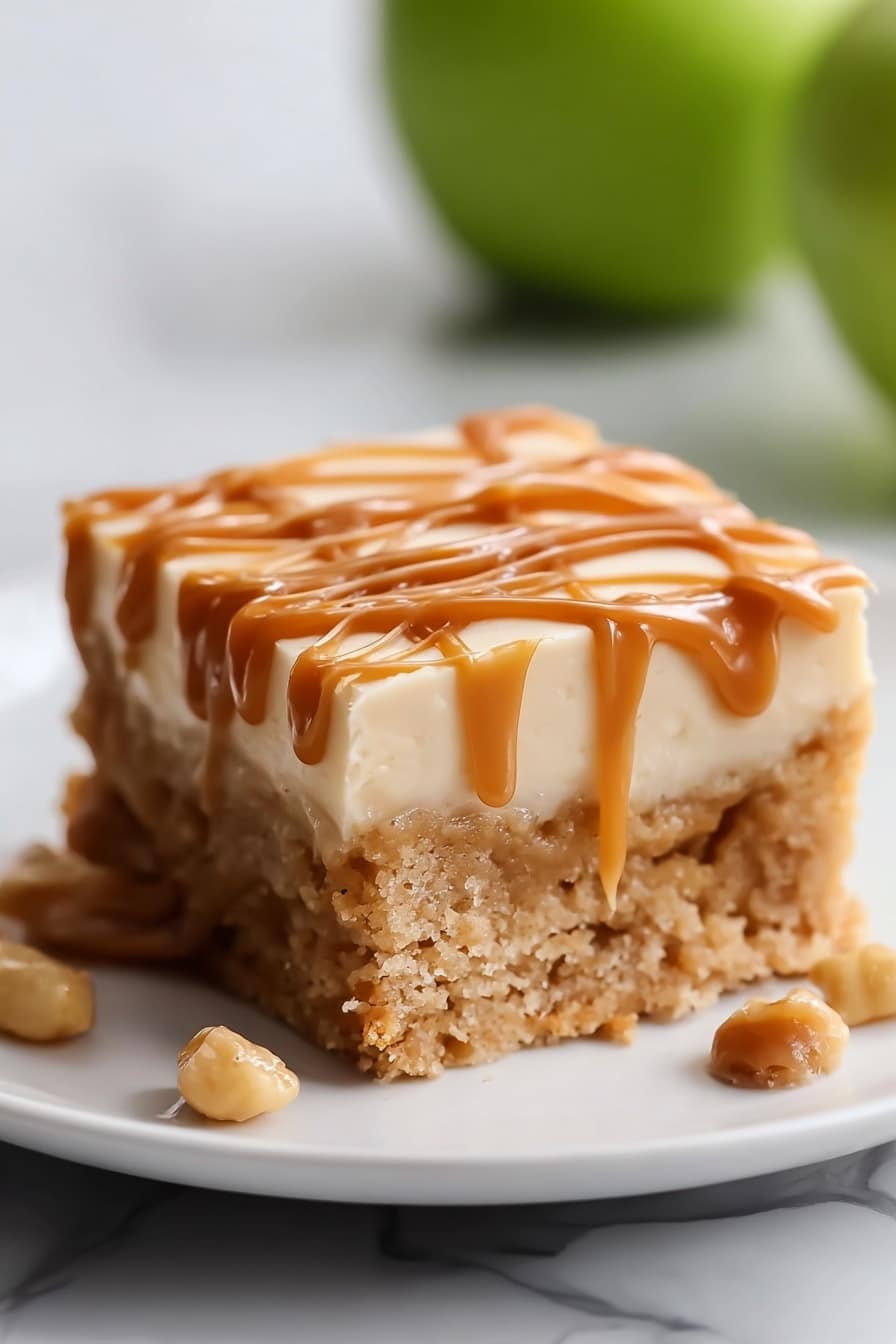 The image shows a single square piece of dessert on a white plate with three visible layers. The bottom layer is a thick, soft, light brown cake with a crumbly texture. The middle layer is a smooth, pale cream frosting, slightly lighter in color than the cake. The top layer is a shiny caramel sauce drizzled in a loose, crisscross pattern that runs slightly down the sides of the cake, adding a glossy texture. There are a few caramel-colored nuts beside the cake on the plate. In the background, a green apple is softly out of focus. The setting has a white marbled texture. Photo taken with an iphone --ar 2:3 --v 7 - Salted Caramel Apple Sheet Cake, fall apple cake, easy apple sheet cake, caramel apple dessert, cozy autumn cake