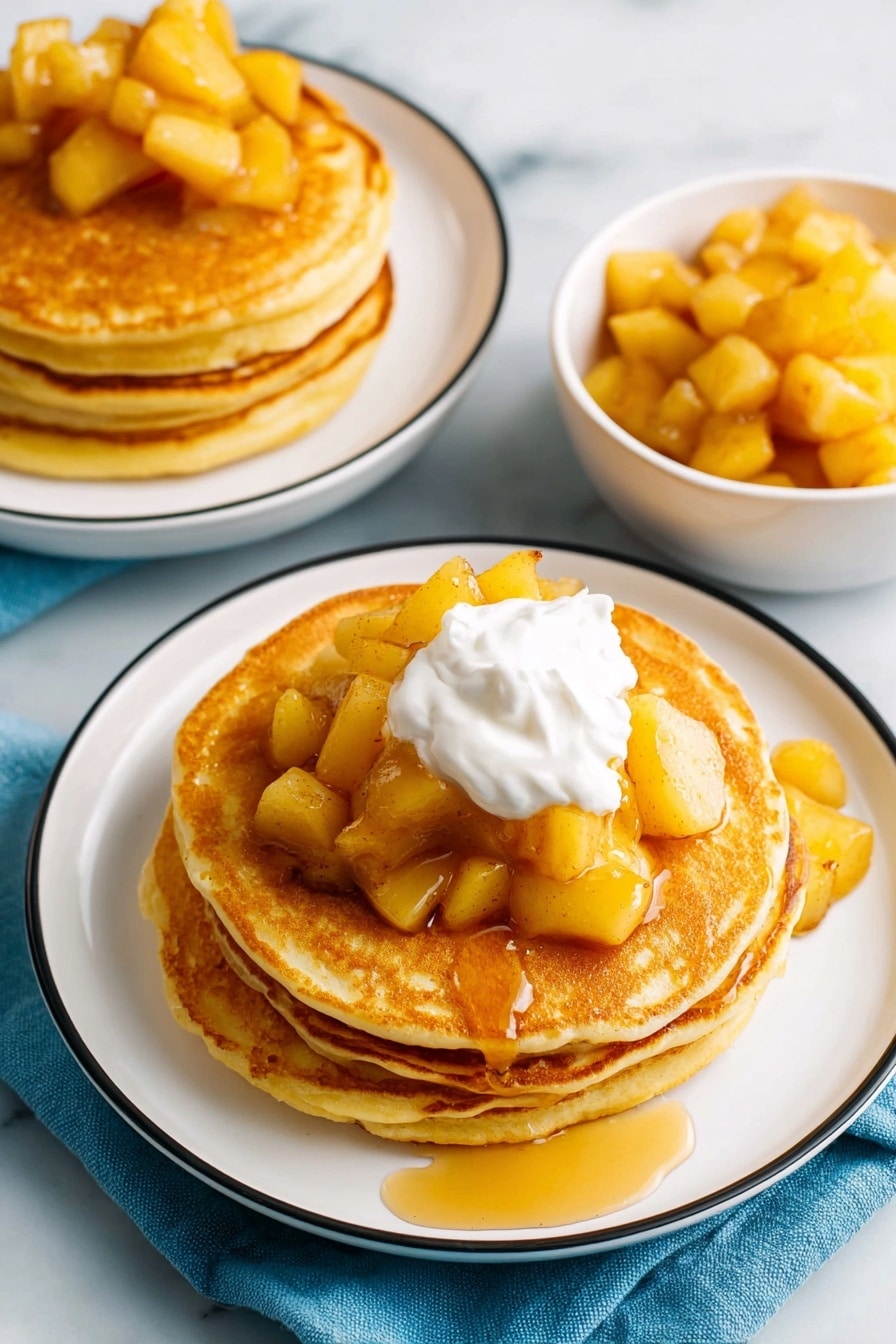 The image shows two stacks of golden brown pancakes on white plates with thin black rims, placed on a white marbled surface. Each stack has about four or five pancakes, with soft, round edges and a smooth texture. The top layer of the pancakes is covered with chunks of cooked, light yellow apple pieces, which have a glossy, syrupy coating that lightly drips over the sides. On the closer plate, a dollop of white whipped cream sits on the center of the top pancake, with honey drizzled over it and the apple pieces. To the right side of the image, there is a white bowl filled with more of the same cooked apple chunks. The setting includes a blue cloth partially visible under the front plate. photo taken with an iphone --ar 2:3 --v 7 - Apple Pancakes, cinnamon apple pancakes, fluffy apple pancakes, easy apple pancake recipe, breakfast with apples