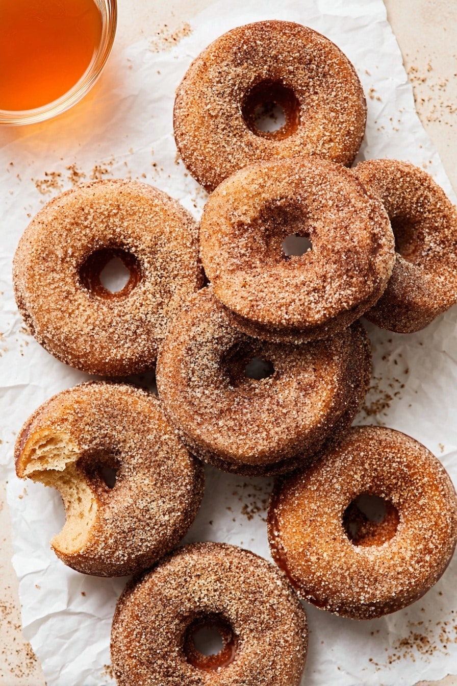 The image shows eight cinnamon sugar doughnuts placed closely on a white marbled textured surface covered with crumpled white parchment paper. The doughnuts are all round with a central hole and evenly coated with a brown sugar and cinnamon mix, giving them a grainy texture on top. Two of the doughnuts have bites taken out, revealing a soft and light brown interior with a slightly coarse crumb. The doughnuts are slightly stacked or leaning on one another, creating a layered look. The top-down view showcases the crumbly sugar coating and soft texture inside, with some sugar granules scattered around the doughnuts. On the upper left corner, a glass cup with orange-colored liquid is partly visible. Photo taken with an iphone --ar 2:3 --v 7 - Gluten Free Apple Cider Donuts, gluten free fall treats, apple cider donut recipe, homemade gluten free donuts, seasonal gluten free desserts