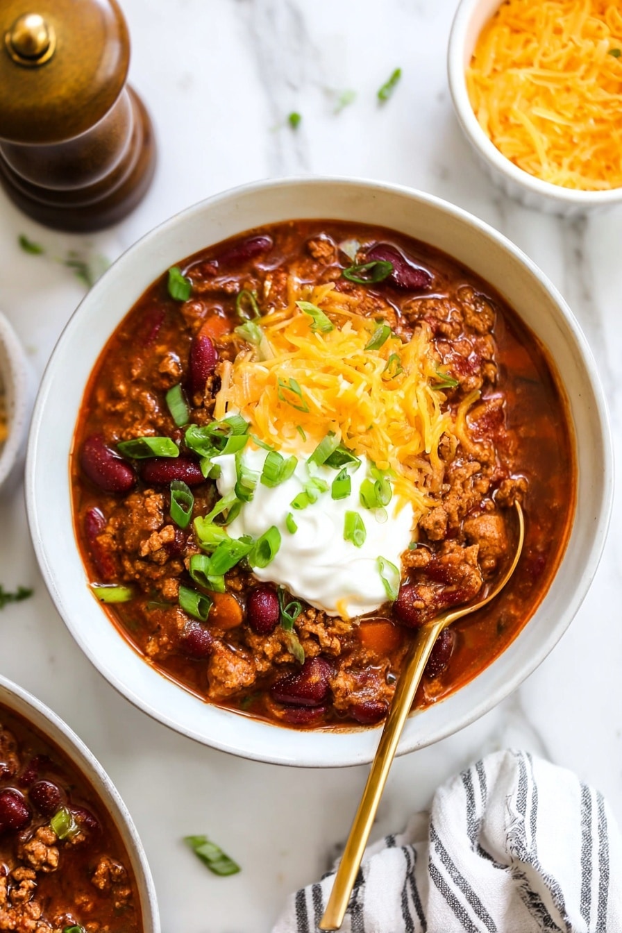 A white bowl filled with thick chili showing three main layers: the bottom layer is rich reddish-brown chili with visible kidney beans and ground meat, the middle layer is a dollop of white sour cream, and the top layer is a mound of shredded bright orange cheddar cheese sprinkled with chopped green onions. A brass spoon is placed inside the bowl on the right side. The bowl sits on a white marbled surface with part of a white bowl of shredded cheddar cheese and another bowl of chili blurred in the background. Photo taken with an iphone --ar 2:3 --v 7 - Classic Chili, best chili recipe, hearty chili, easy chili recipe, flavorful chili