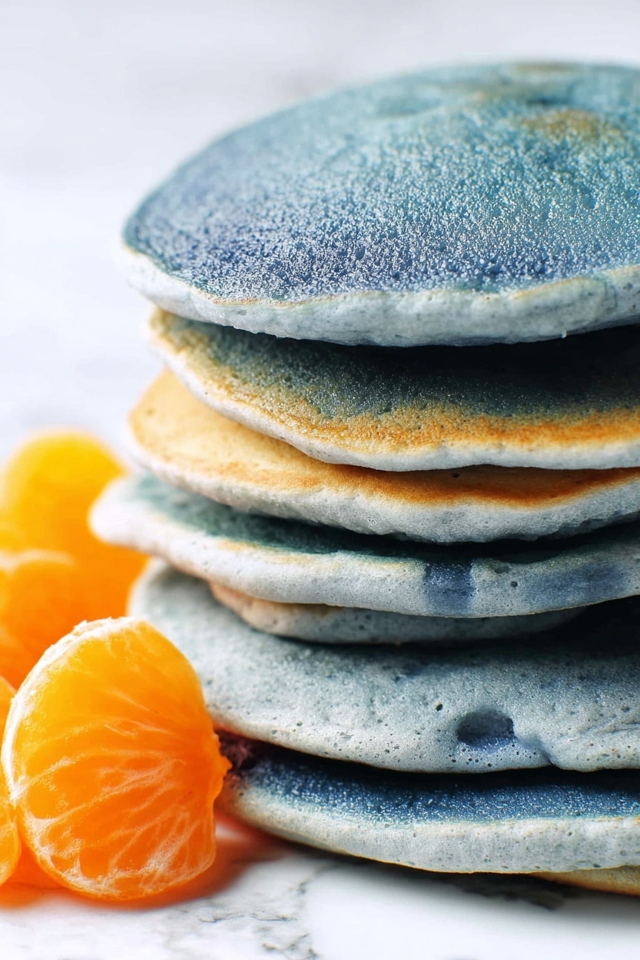 A stack of six thick, round pancakes with a soft and slightly uneven texture, each pancake showing a blue-grey color with soft gradients and subtle cracks along the edges. The pancakes are closely stacked, showing the layers clearly from the side. On the left, there is a small portion of bright orange fruit, possibly tangerine segments, placed on a white marbled surface. The background is softly blurred with various white tones, focusing attention on the stack. photo taken with an iphone --ar 2:3 --v 7 - Blue Pancakes No Artificial Coloring, natural blue pancakes with spirulina, healthy vibrant pancake recipe, quick homemade blue breakfast, egg-free blue pancake ideas