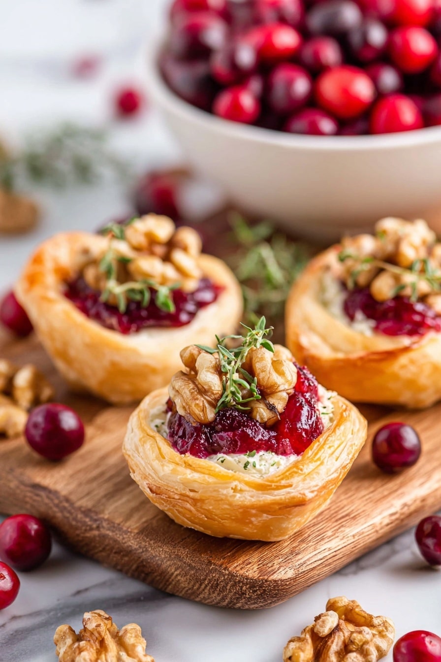 The image shows small puff pastry cups with three layers; the first layer is a golden, flaky pastry forming the shell, the second layer inside is a light cream cheese base, and the third layer is a bright red cranberry sauce topped with chopped walnuts and fresh green thyme leaves. The pastry cups sit on a wooden board with scattered fresh red and dark red cranberries and some walnut pieces around. In the background, there is a white bowl filled with fresh cranberries, all placed on a white marbled surface. Photo taken with an iphone --ar 2:3 --v 7 - Cranberry Brie Bites, Cranberry Brie Bites appetizer, easy cranberry brie bites, festive puff pastry appetizers, holiday cranberry brie bites