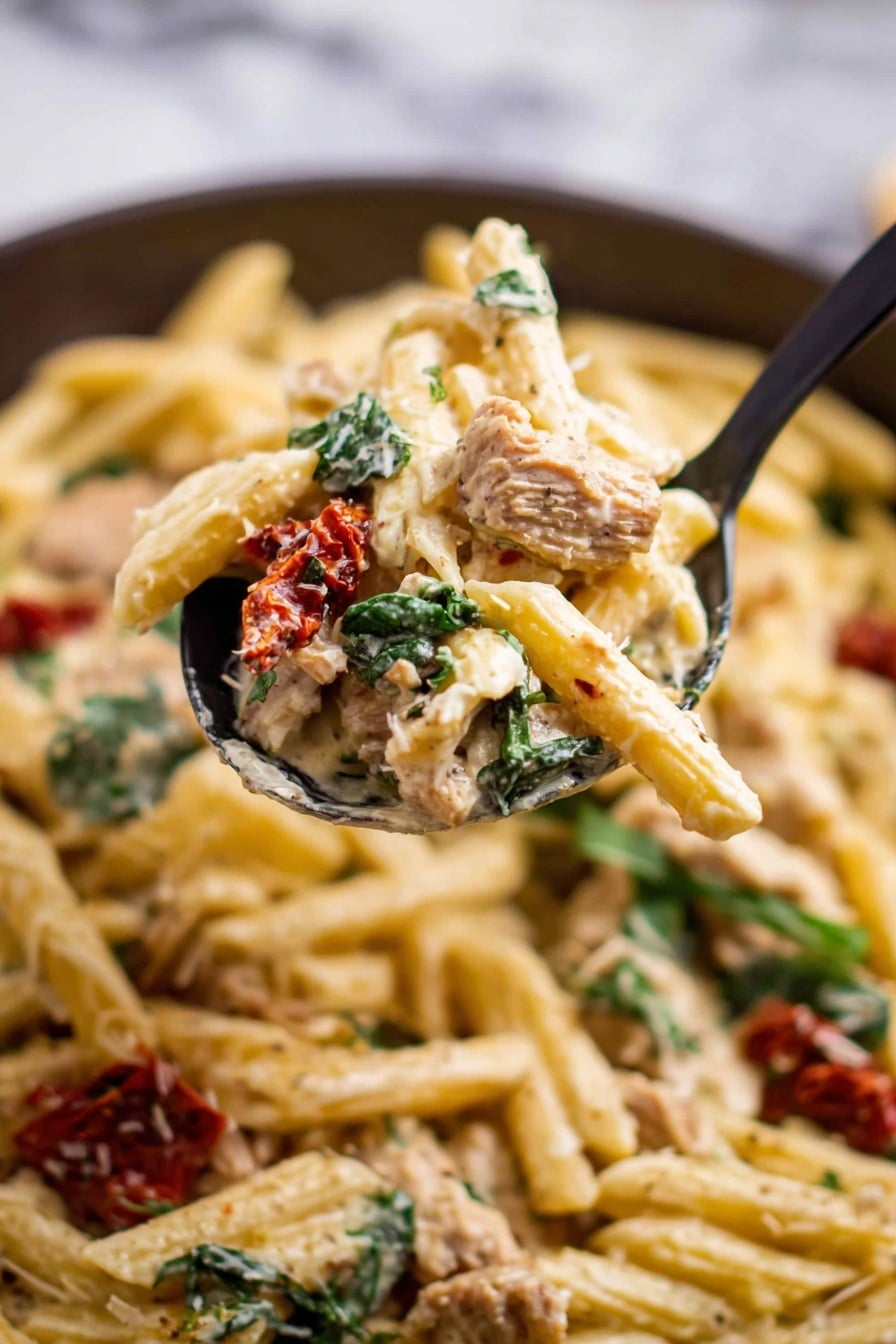 The image shows a close-up of a spoon lifting a creamy pasta dish, with the background filled with the same pasta. The dish has three main layers: the pasta which is pale yellow with a smooth and soft texture, pieces of light brown meat scattered throughout, and dark green leafy herbs adding a fresh contrast. There are also small bits of red sun-dried tomatoes adding another color and texture. The spoon is black and holds a mix of all the parts, showing a thick creamy sauce coating the pasta and other ingredients. The background surface is a white marbled texture. Photo taken with an iphone --ar 2:3 --v 7 - One Pot Italian Chicken Pasta, Italian chicken pasta, easy one pot pasta, creamy chicken pasta, quick weeknight dinner