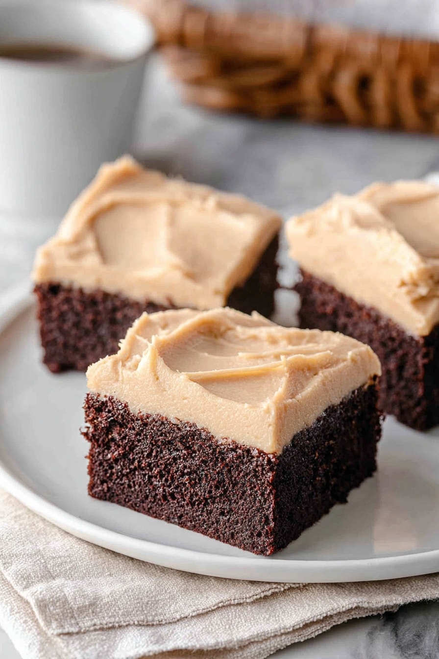 Three square pieces of dark brown chocolate cake with a thick, smooth light brown frosting layer on top are placed on a white plate. The frosting has a creamy, slightly swirled texture. The cake looks moist and dense. The plate sits on a white marbled surface with a soft background showing a basket and a white cup. Photo taken with an iphone --ar 2:3 --v 7 - Frosted Coffee Brownies, coffee-flavored brownies, chocolate coffee dessert, easy coffee brownies, chocolate brownie recipes