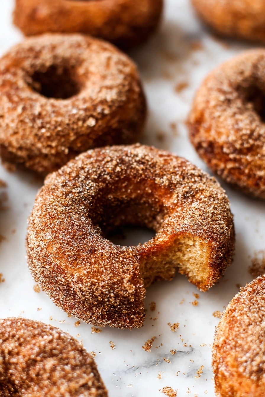 The image shows several round donuts covered with a layer of rough sugar and cinnamon crystals. Each donut has a golden brown color with darker shades blending with the sugar coating, giving a crispy texture look. The donuts are arranged close together on a white marbled surface with small crumbs scattered around. One donut is broken with a bite missing, revealing a soft, textured inner layer. The focus is on the front donut while the others blur softly in the background. photo taken with an iphone --ar 2:3 --v 7 - Gluten Free Apple Cider Donuts, gluten free fall treats, apple cider donut recipe, homemade gluten free donuts, seasonal gluten free desserts