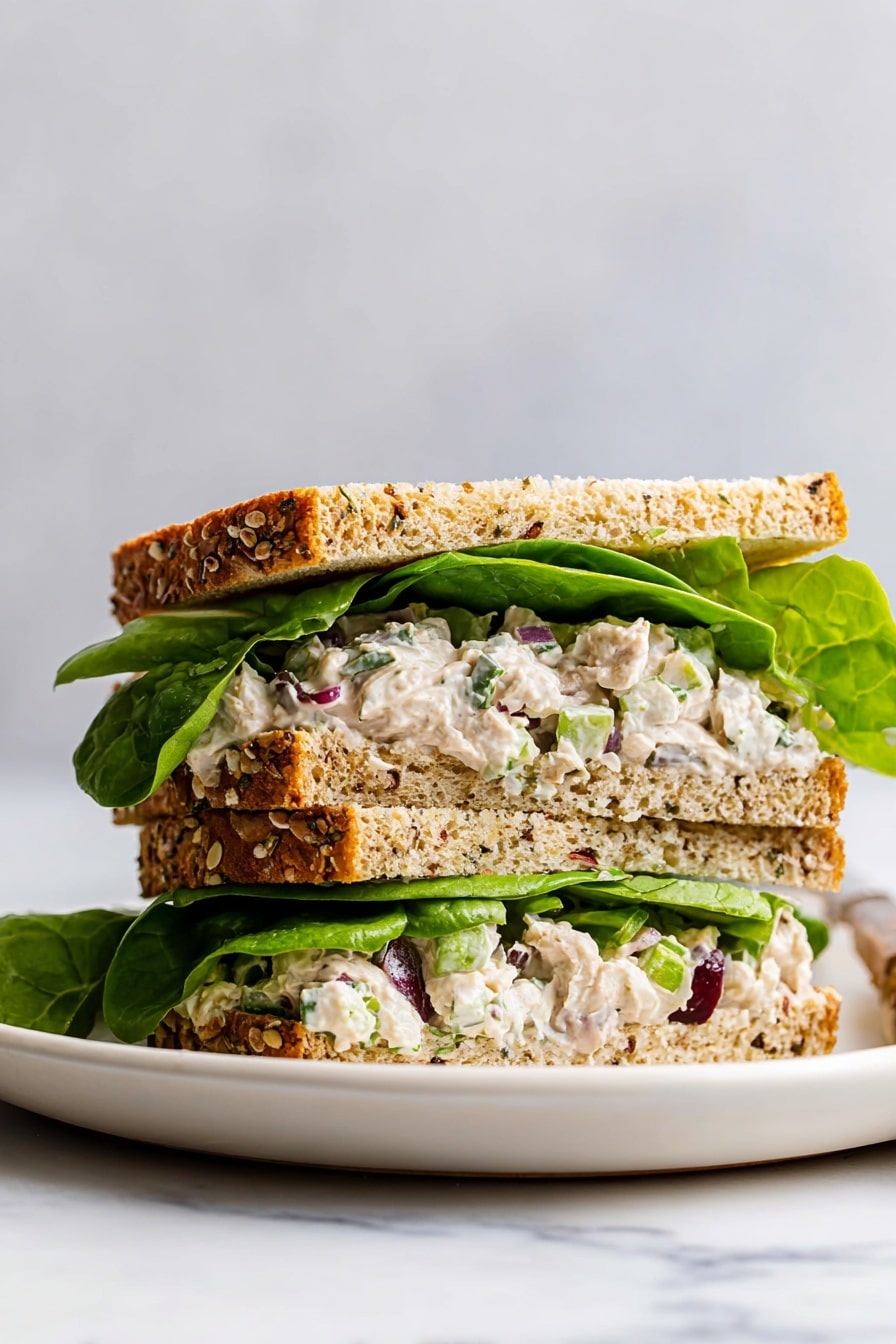 A sandwich is shown on a white plate with a white marbled background. The sandwich has three visible layers: the bottom and top layers are thick slices of multigrain bread with seeds and a golden-brown crust. Inside, there are two layers of fresh green lettuce leaves; one layer sits directly on the bottom slice and the other layer is below the top slice. Between the lettuce layers, there is a creamy chicken salad mixed with small pieces of red onion and celery, creating a textured mix of white, purple, and green colors. Photo taken with an iphone --ar 2:3 --v 7 - Leftover Turkey Salad, easy leftover turkey ideas, cranberry turkey salad, quick turkey salad recipe, turkey leftovers ideas