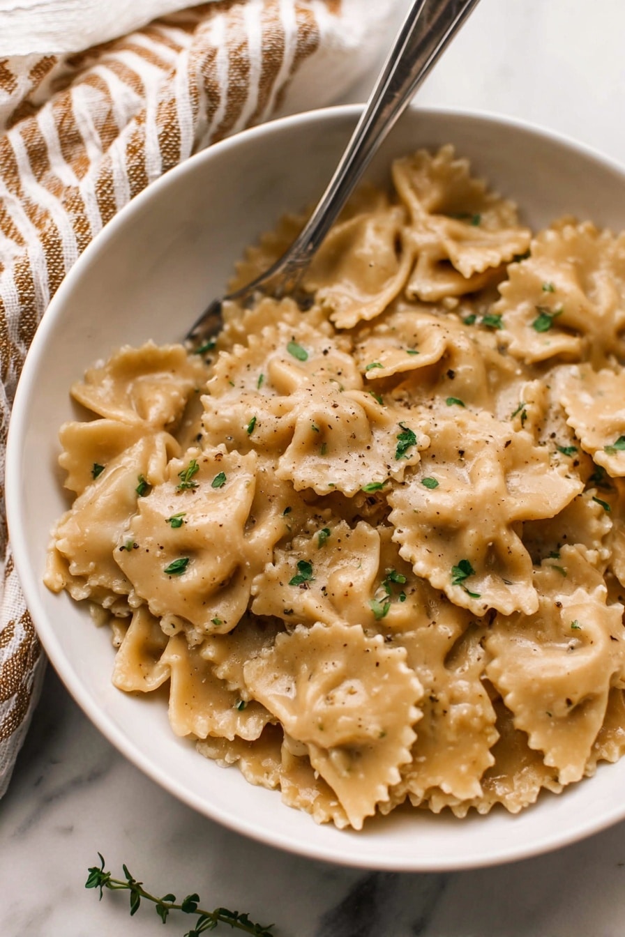 A white bowl filled with soft, folded pasta pieces coated in a light brown creamy sauce, with tiny green herb sprigs scattered on top. The pasta has wavy edges and a slightly rough texture. A silver fork rests inside the bowl, leaning against the side. The background shows a beige and brown striped cloth on a white marbled surface. Photo taken with an iphone --ar 2:3 --v 7 - French Onion Pasta, French Onion Soup Pasta, Caramelized Onion Pasta, Cozy Pasta Recipes, Easy Pasta Dinner