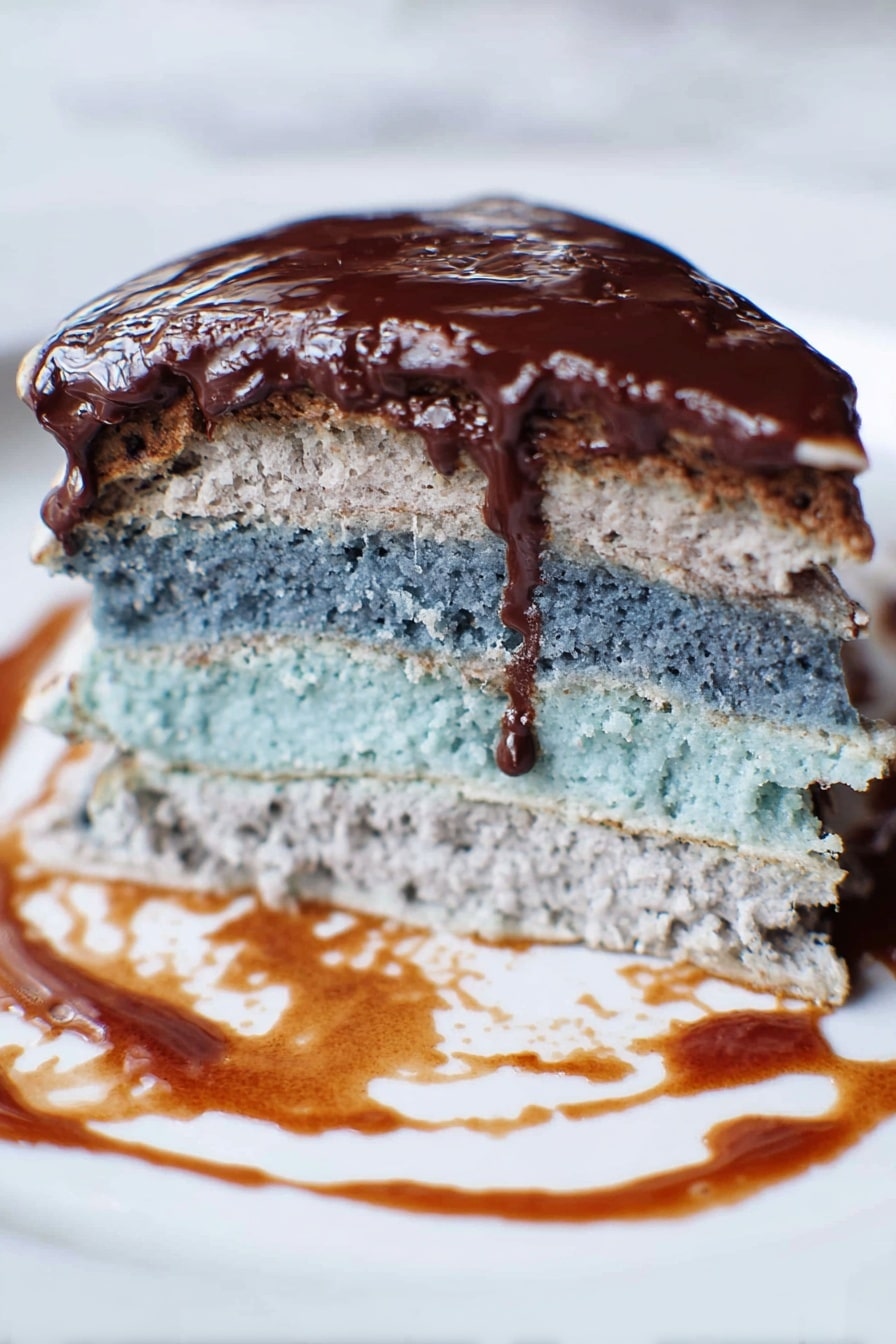 The image shows a close-up of a slice of layered pancakes on a white plate with a white marbled texture surface beneath. The bottom layer is thick and light gray with a soft, crumbly texture. Above it is a thinner, light blue layer, which also looks soft but a bit more dense. The top layer is a darker shade of brown, covered in shiny, smooth chocolate sauce that drips slightly over the sides. There are some smears of chocolate sauce on the plate around the pancakes, showing a bit of messiness. The lighting highlights the moist texture and glossy topping of the top layer. photo taken with an iphone --ar 2:3 --v 7 - Blue Pancakes No Artificial Coloring, natural blue pancakes with spirulina, healthy vibrant pancake recipe, quick homemade blue breakfast, egg-free blue pancake ideas