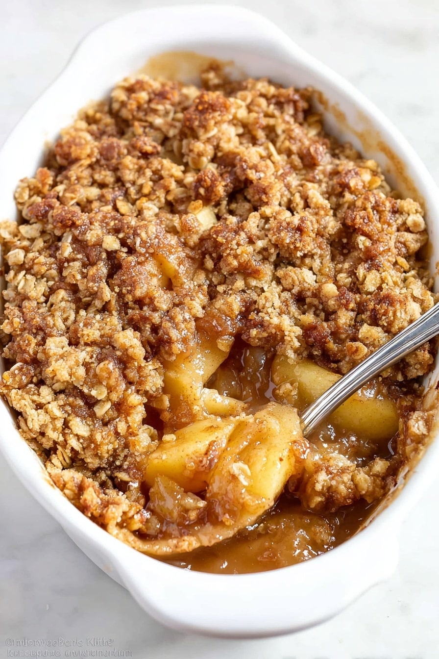 The image shows a white oval dish filled with a warm apple crumble. The bottom layer is made of soft, cooked apple slices in a shiny, sticky golden-brown sauce. On top, there is a thick, rough layer of crumbly oat topping with a mix of small and medium brown pieces, creating a textured surface. The crumble topping has a golden color with some darker brown spots from baking. A metal spoon is partly dipping into the crumble from the right side, revealing the soft apple filling beneath in the center. The dish is placed on a smooth white marbled surface. photo taken with an iphone --ar 2:3 --v 7 - The Best Apple Crisp, apple crisp dessert, cinnamon apple crisp, easy apple crisp, comforting fall desserts