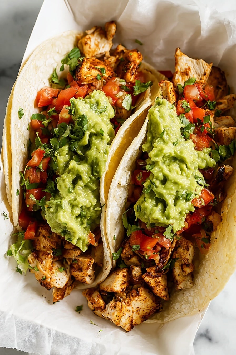 Two soft corn tortillas lie side by side on crumpled white paper over a white marbled surface. Each tortilla holds a layer of cooked, light brown chicken pieces with slight char marks, topped with a colorful salsa mix of diced red tomatoes and green herbs. A generous scoop of chunky, bright green guacamole sits on top of the salsa in each taco. In the background, a white bowl of guacamole and a white bowl with chopped green herbs are partially visible. Photo taken with an iphone --ar 2:3 --v 7 - Chicken Tacos with Homemade Marinade, homemade chicken taco marinade, easy chicken taco recipe, flavorful chicken tacos, quick dinner tacos