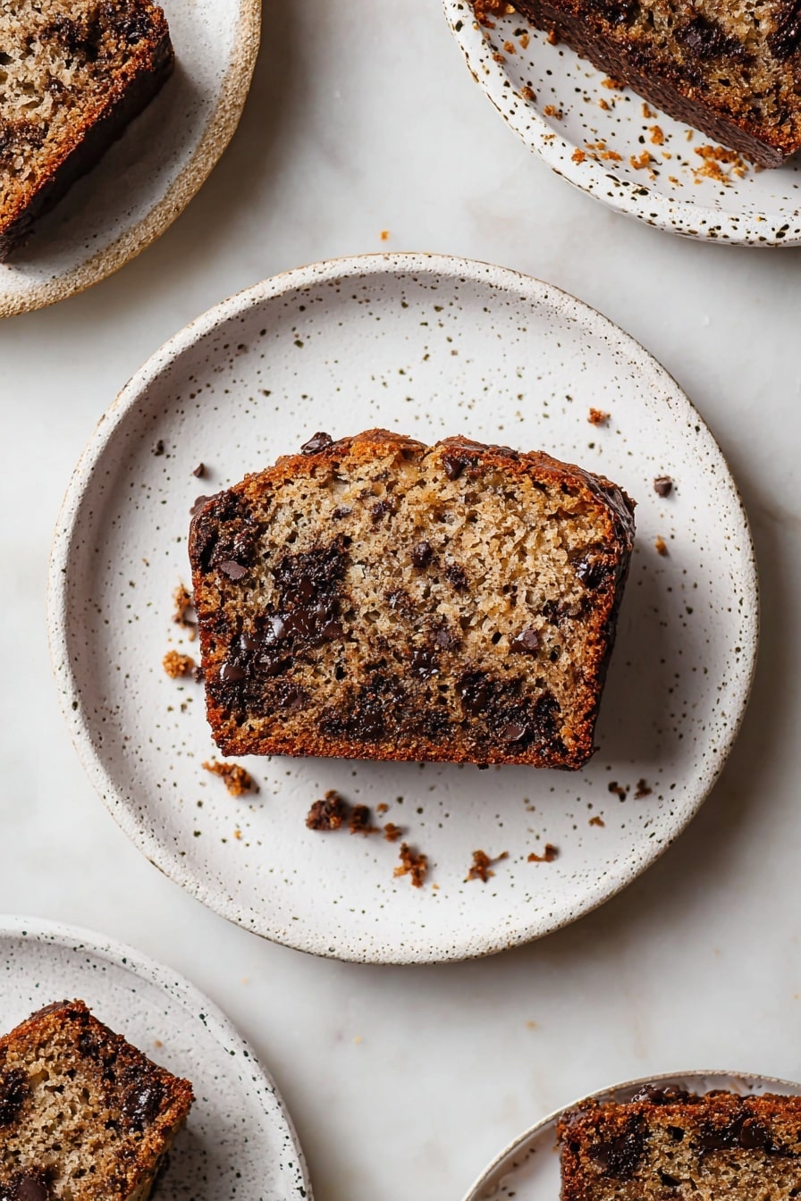 A single slice of moist banana bread sits in the center of a white speckled plate. The bread has a rich golden-brown crust and a soft, light brown inside, dotted generously with melted dark chocolate chips that create small, shiny, uneven patches throughout. Small crumbs are scattered around the slice on the plate, showing a slightly crumbly texture. Other white speckled plates with similar pieces of banana bread and crumbs surround the main plate, all set over a white marbled background. photo taken with an iphone --ar 2:3 --v 7 - Gluten-Free Banana Bread with Chocolate Chips, gluten-free banana bread recipe, healthy banana bread with chocolate, moist gluten-free banana loaf, easy gluten-free banana bread