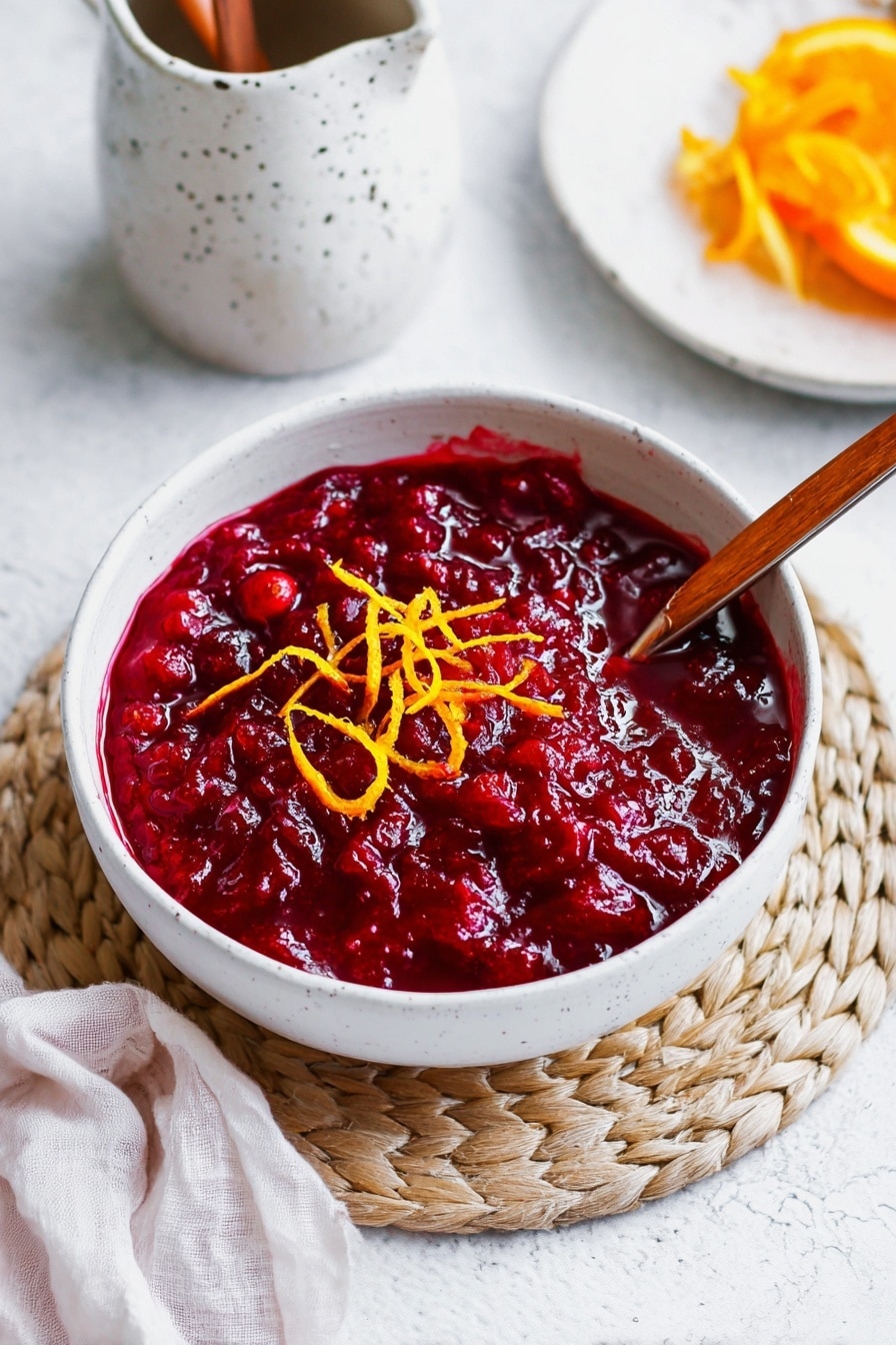 A white bowl filled with chunky deep red cranberry sauce with a shiny, thick texture sits in the center on a round braided natural-colored mat. On top of the sauce are thin orange peel strips adding a pop of bright color. A wooden spoon with a light brown color rests partly in the bowl, touching the sauce. The bowl is placed on a white marbled surface. Nearby, a white ceramic container with speckles and a wooden spoon rests above the bowl, and on the right side, parts of a white plate with more orange peel strips can be seen. A soft white cloth is draped on the bottom left corner. Photo taken with an iphone --ar 2:3 --v 7 - Cranberry Orange Sauce, holiday cranberry sauce, citrus cranberry side, easy cranberry sauce, fresh fruit cranberry recipe