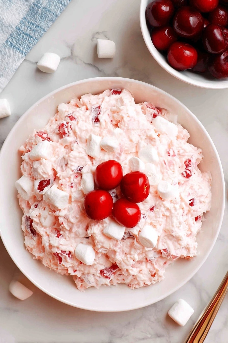 The dish is served on a white plate filled with a creamy, fluffy salad that is pale pink with white marshmallow pieces mixed evenly throughout. The texture looks soft and slightly chunky, with visible bits of fruit or cherry embedded in the mixture. In the center, there is a small pile of bright red whole and halved cherries that add a splash of color and contrast. The plate sits on a white marbled surface next to a white bowl filled with more cherries and small white marshmallows scattered nearby. The overall look is light, creamy, and inviting, with a mix of smooth, chunky, and juicy textures. Photo taken with an iphone --ar 2:3 --v 7 - Cranberry Fluff, Cranberry Fluff dessert, holiday cranberry salad, creamy cranberry fluff, easy cranberry fluff recipe