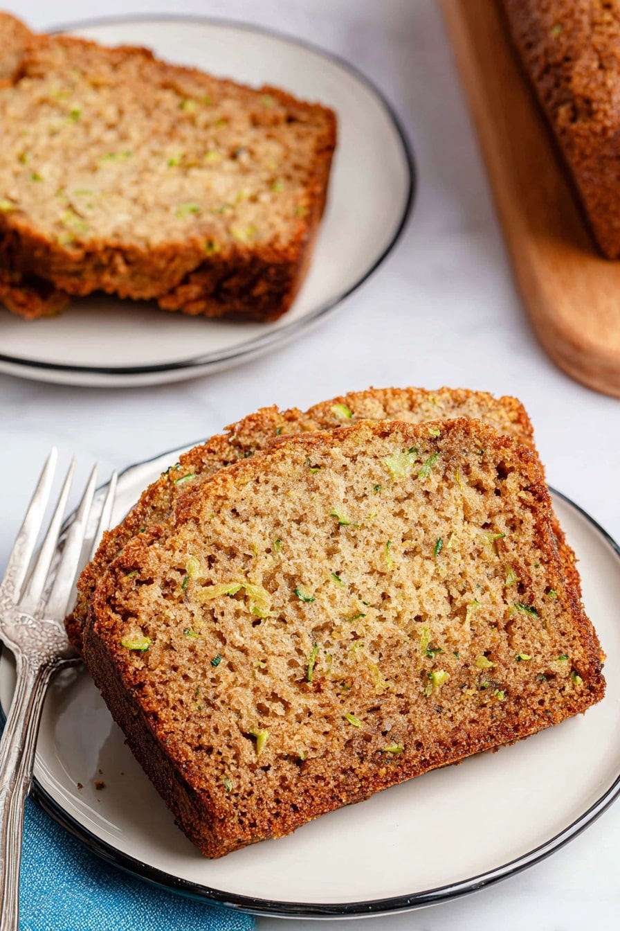 A close-up view shows a single thick slice of moist, golden-brown zucchini bread with a slightly rough texture and visible green flecks of zucchini throughout the soft interior. The slice rests on a white plate with a thin black rim, accompanied by a silver fork on the left side, near a blue cloth napkin. In the background, there is a matching white plate holding another slice and part of the remaining loaf sits on a wooden cutting board, all placed on a white marbled surface. The bread has a well-baked crust with darker edges and a visibly tender crumb inside. photo taken with an iphone --ar 2:3 --v 7 - Banana Zucchini Bread, moist zucchini bread recipe, healthy banana zucchini bread, easy zucchini bread with bananas, quick banana zucchini loaf