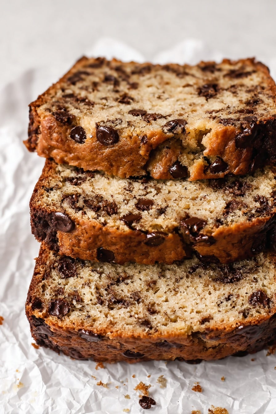 The image shows three thick slices of chocolate chip banana bread stacked on top of each other. Each slice has a light brown crumb with a moist, slightly crumbly texture, sprinkled with dark brown melted chocolate chips evenly spread throughout. The crust is darker brown, firm, and slightly shiny, wrapping the edges of each slice. The bread rests on white crinkled parchment paper set against a white marbled textured background. photo taken with an iphone --ar 2:3 --v 7 - Protein Banana Bread with Chocolate Chips, healthy banana bread, high-protein banana bread, nutritious chocolate chip banana loaf, easy protein banana bread recipe