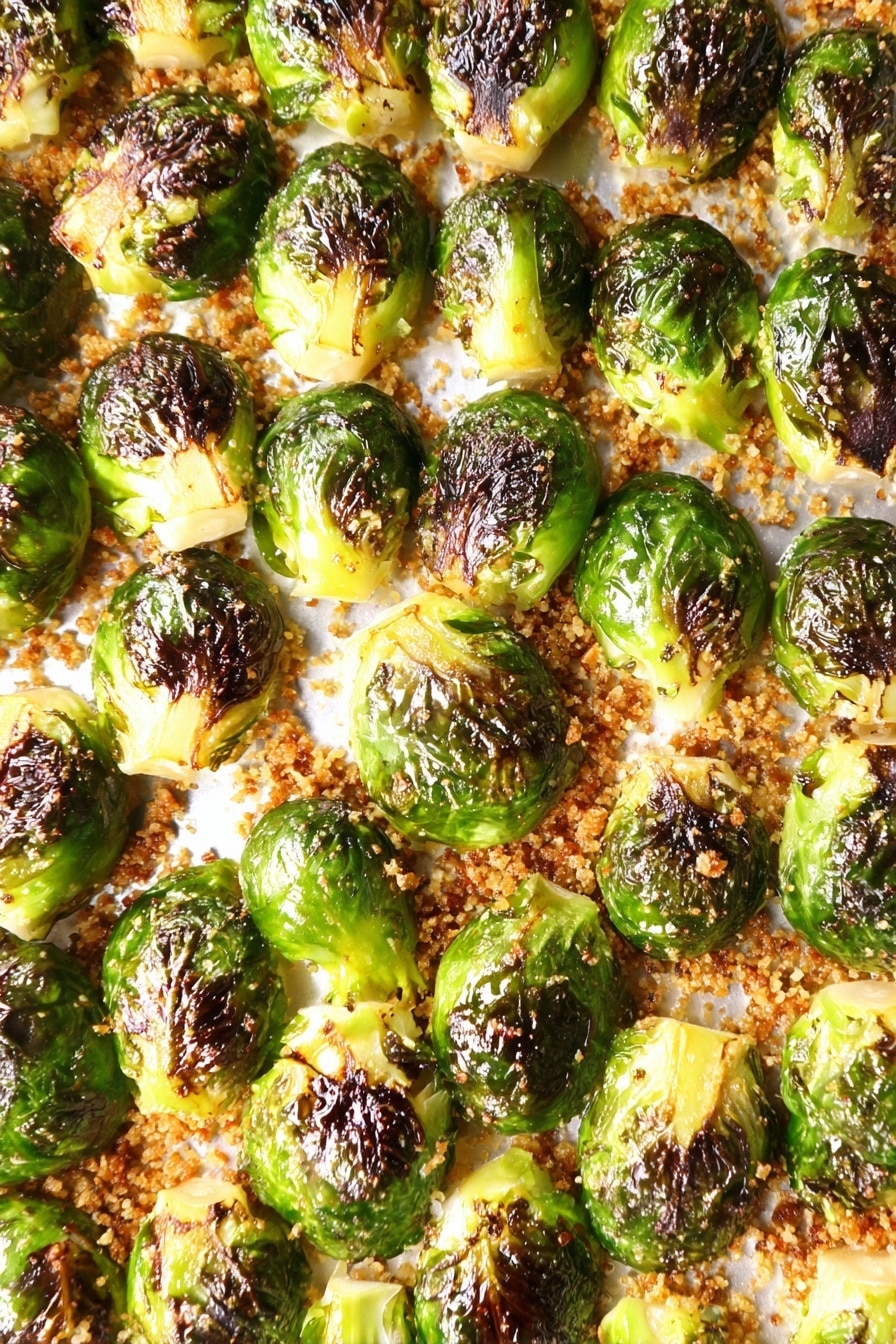 The image shows a tray full of halved Brussels sprouts arranged in tight rows, with vibrant green outer leaves and dark brown charred spots on the top, giving a crispy look. The sprouts sit on a golden-brown, slightly crunchy textured base that looks like seasoned crumbs spread evenly across the white tray surface. The contrast between the bright green and dark toasted colors highlights the roasted effect on the vegetable halves. photo taken with an iphone --ar 2:3 --v 7 - Oven Roasted Parmesan Brussel Sprouts, roasted Brussels sprouts with Parmesan, easy Brussels sprouts recipe, healthy roasted Brussels sprouts, crispy Brussel Sprouts with Parmesan