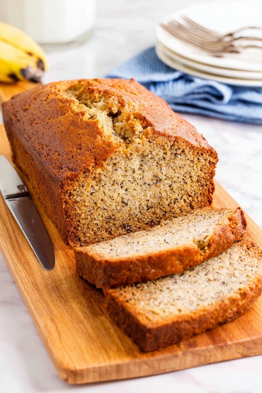 A loaf of banana bread with a golden brown crust sits on a wooden cutting board, sliced to show the soft, moist inside filled with tiny dark specks of banana and nuts. The bread has a rough, textured top with a deep crack running through the middle. To the left of the bread is a silver knife resting on the board. In the background, there are white plates stacked with silver forks on a blue cloth, all set on a white marbled surface. photo taken with an iphone --ar 2:3 --v 7 - One-Bowl Banana Bread, Easy Banana Bread, Moist Banana Bread, Quick Banana Bread Recipe, Simple Banana Bread