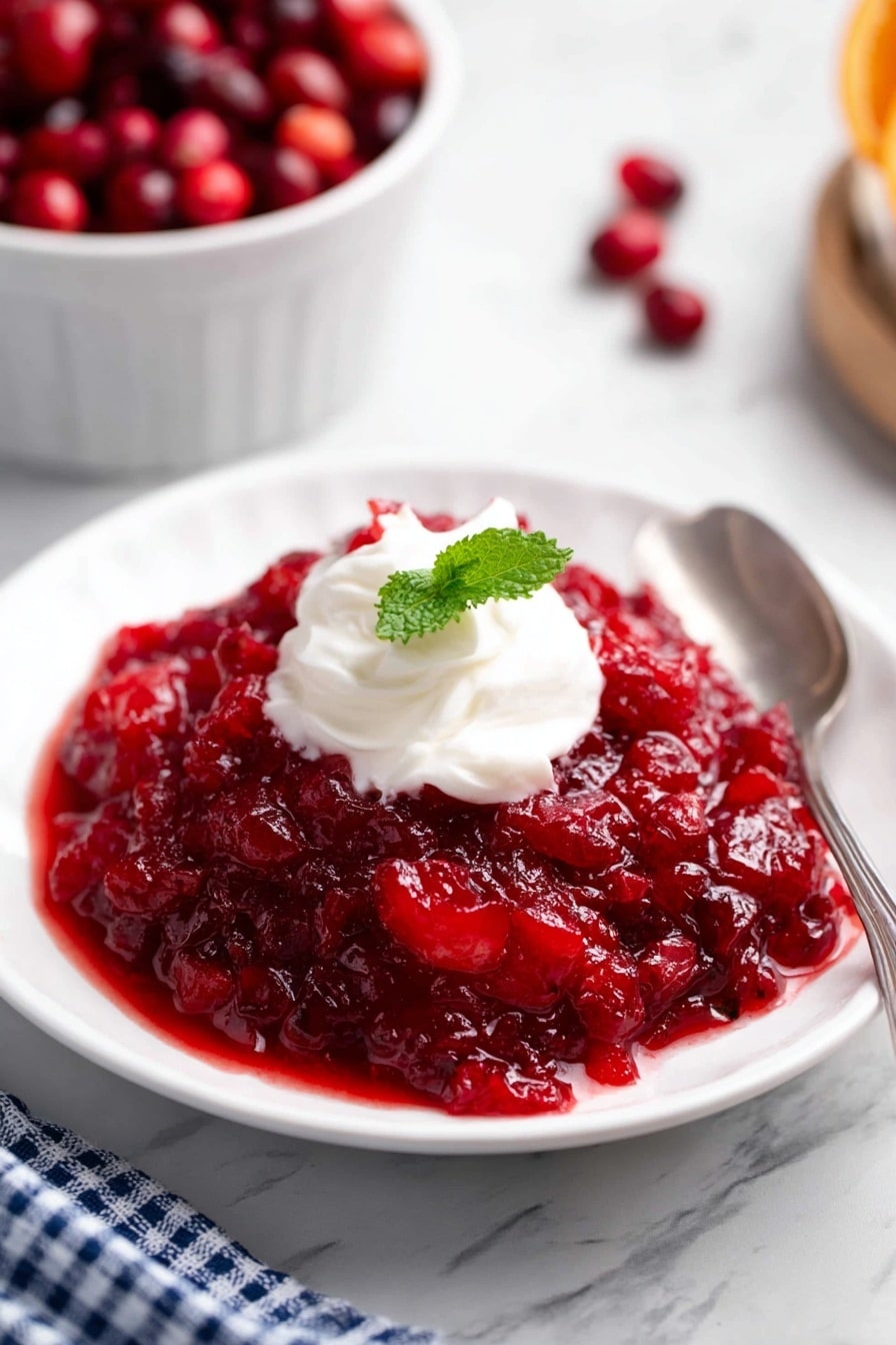 A white plate has one main layer of chunky bright red cranberry sauce with visible soft fruit pieces. On top in the center is a small swirl of white cream with a smooth texture, and beside the cream is a single green mint leaf. A silver spoon rests on the right edge of the plate. The plate is set on a white marbled surface, with a white bowl filled with whole red cranberries blurred in the background. A blue and white checkered cloth is partly visible near the bottom edge. Photo taken with an iphone --ar 2:3 --v 7 - Cranberry Jello Salad, Cranberry Jello Salad Ingredients, Festive Cranberry Salad, Holiday Jello Salad, Easy Cranberry Salad